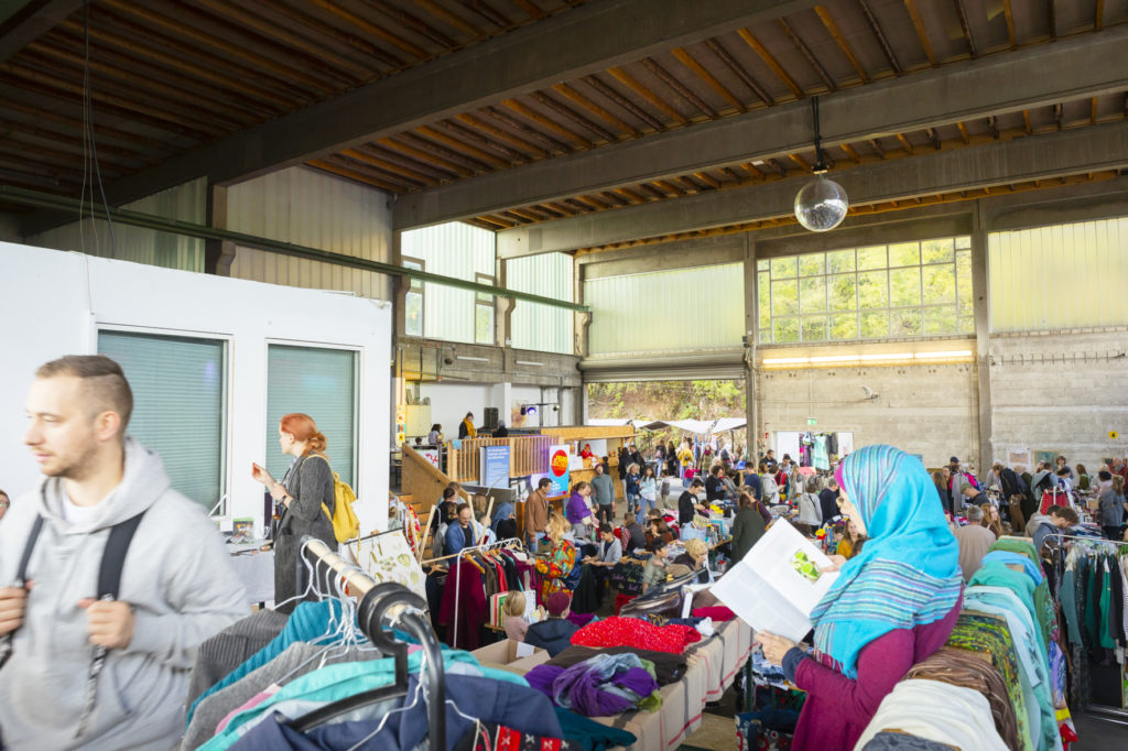 Ein Gewusel von Menschen in einer Industriehalle, die sich durch die engen Gänge des Trödelmarktes drängen. Im Vordergrund ist ein Stand zu sehen der Kleidung verkauft und eine Frau, die in einem Buch liest